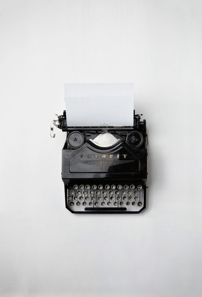 old style typewriter viewed from above on a white surface. Contrast of black and white. A sheet of blank paper waits. 